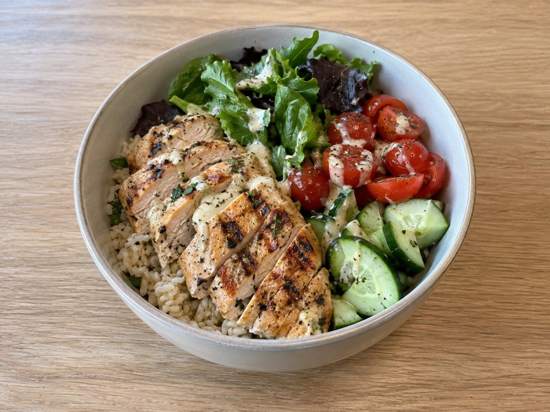 Lunch bowl with sliced grilled chicken, rice, leafy greens, tomatoes, and cucumber.